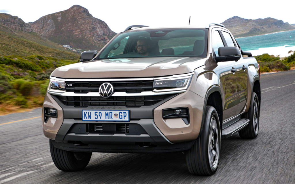 Na foto há o carro VW Amarok 2024 de cor marrom, em uma avenida, ao fundo da imagem é possível ver uma praia